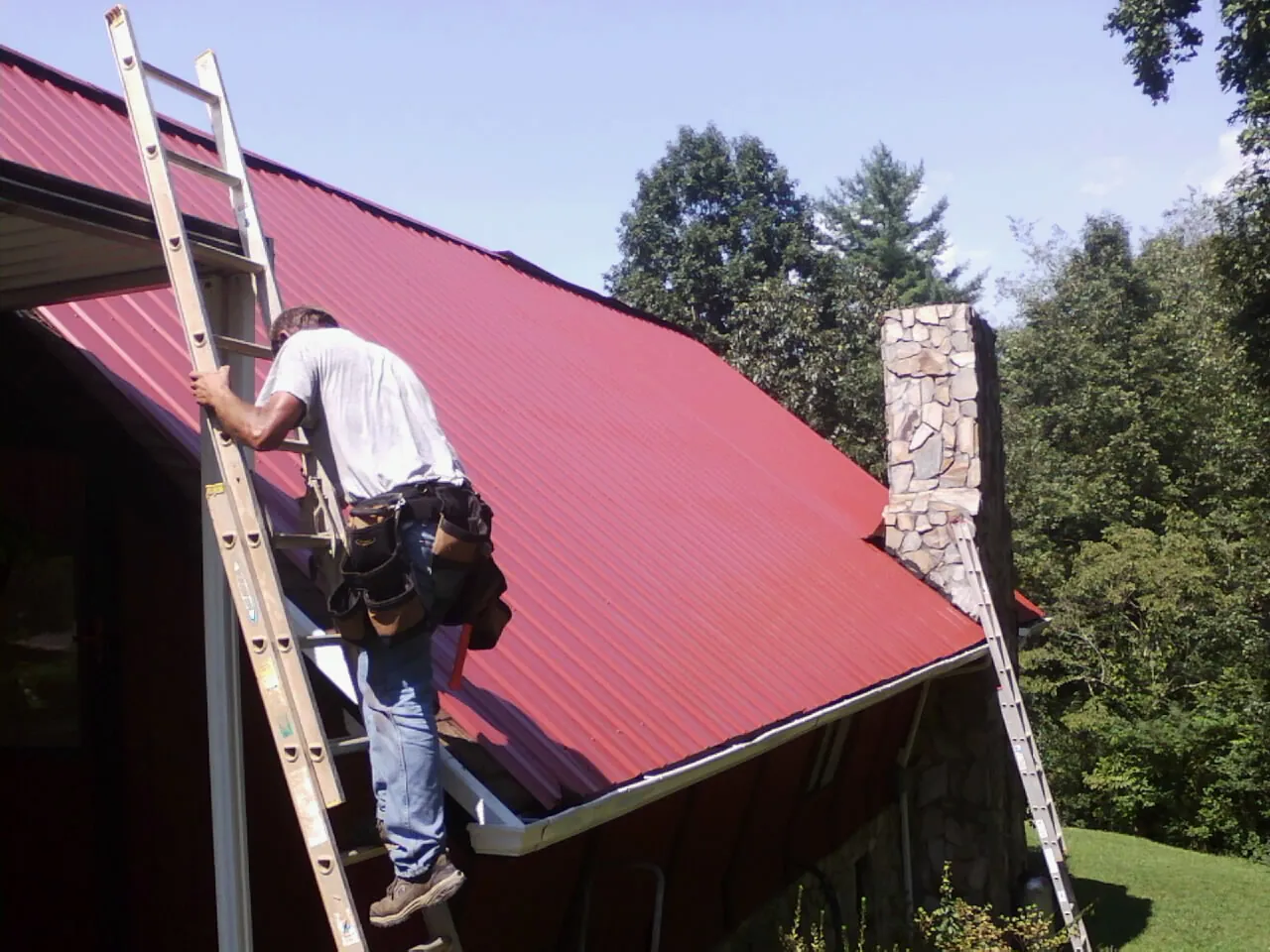 Roofer on a ladder beside a red metal roof and stone chimney — Soffit & Fascia Repair by Roofing Brothers in Lowell